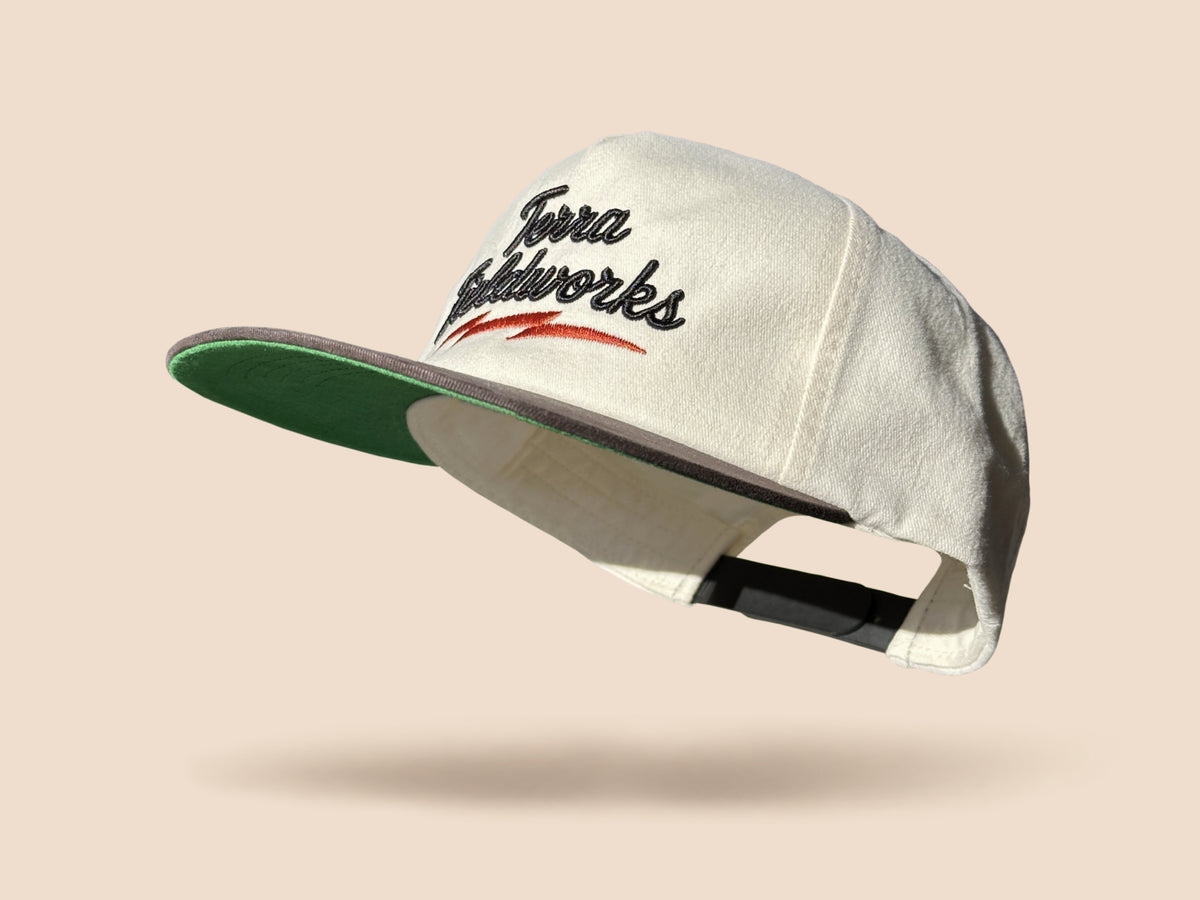 Lightning Bolt Snapback Hat in cream/black with black puff embroidered Terra Fieldworks script and orange lightning bolt showing the green underbill